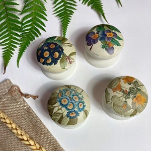 May include: Four round wooden knobs with floral designs. The knobs are white with a floral pattern in shades of blue, yellow, and green. The knobs are arranged in a square shape on a white surface.