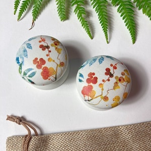 May include: Two small, round, wooden knobs with a white background and a floral pattern in shades of orange, yellow, blue, and green. The knobs are sitting on a white surface next to a small hessian bag.