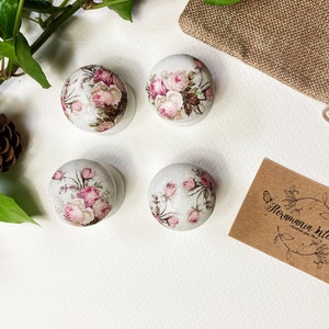 May include: Four white round knobs with floral designs featuring pink roses. The knobs are displayed on a white surface with a brown hessian bag and a business card that reads "Flormania Interiors".