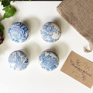 May include: Four small round ceramic boxes with blue and white floral designs. The boxes are arranged in a square pattern on a white surface. A brown paper card with the text "Hormannia Interiors" is in the lower right corner of the image.