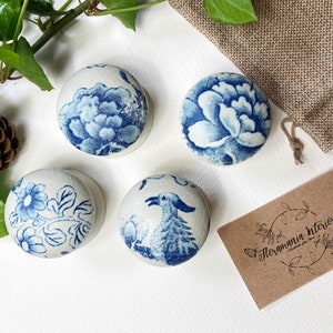 Può includere: Quattro pomelli in ceramica bianca con disegni floreali blu e bianchi. I pomelli presentano diversi motivi floreali, tra cui un uccello e un grande fiore. I pomelli sono disposti su una superficie bianca con un piccolo cono di pino e foglie verdi sullo sfondo. Il testo "Floramania interiors" è stampato su un cartoncino marrone.