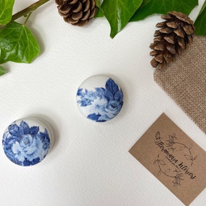 Op de afbeelding: Twee kleine ronde keramische knoppen met een blauw en wit bloemenpatroon. De knoppen bevinden zich op een witte achtergrond met een bruin papieren etiket waarop "floramania hilhorst" staat.