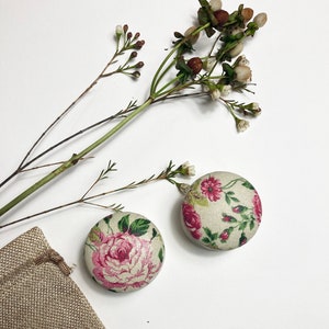 Può includere: Due bottoni rotondi rivestiti in tessuto con un motivo floreale. I bottoni sono rosa e bianchi con un motivo floreale. I bottoni sono su uno sfondo bianco.