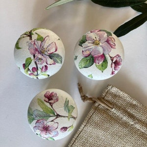 Può includere: Tre pomelli rotondi in legno con sfondo bianco e un design floreale con fiori rosa e bianchi con foglie verdi. I pomelli sono esposti su una superficie bianca con un sacco di juta sullo sfondo.