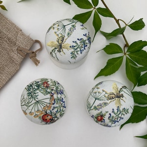 May include: Three round ceramic containers with floral and insect designs. The containers feature a white background with a colorful floral pattern and images of bees and ladybugs.