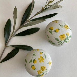 Op de afbeelding: Twee ronde witte knoppen met een geel bloemenpatroon. De knoppen liggen op een wit oppervlak met een groene olijftak.