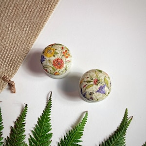 May include: Two round, white ceramic knobs with a floral design. The knobs feature a variety of flowers in shades of yellow, red, purple, and green. The knobs are on a white background.