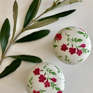 May include: Two white round knobs with a floral pattern. The knobs have a white background with red flowers and green leaves.
