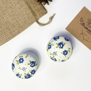 Può includere: Due pomelli in ceramica bianca con un motivo floreale blu e verde. I pomelli sono rotondi e hanno una superficie liscia.