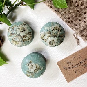 Może przedstawiać: Trzy okrągłe, jasnoniebieskie gałki do szafek z kwiatowym wzorem w kolorze białym i brązowym. Gałki ułożone są na białej powierzchni z małym workiem jutowym, szyszką i gałązką bluszczu. Obecna jest również wizytówka.