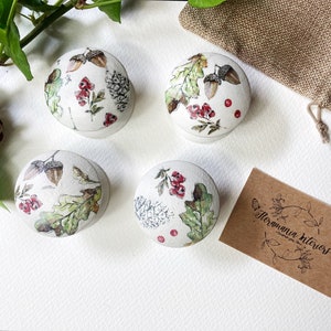 May include: Four small round white boxes with lids, each decorated with a different autumnal design featuring acorns, leaves, and berries. The boxes are arranged in a square on a white surface. A small brown paper card with the text "Floramania Interiors" is in the lower right corner of the image.