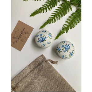 Może przedstawiać: Dwie małe, okrągłe, białe pudełeczka ceramiczne z niebieskimi wzorami kwiatów niezapominajek. Pudełeczka stoją na białym tle, obok brązowego worka jutowego i zielonych liści paproci. Na brązowej kartce widnieje napis "Помнишь лето".