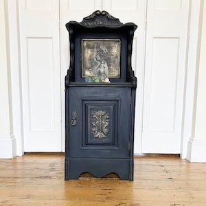 May include: A black painted wooden cabinet with a single door and a shelf. The cabinet has a decorative carved design on the door and a gold-toned frame around the shelf. The shelf has a vintage-style print inside.