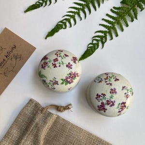 Op de afbeelding: Twee kleine ronde doosjes met een bloemenpatroon. De doosjes zijn wit met paarse bloemen en groene bladeren. De doosjes staan op een bruine jute ondergrond.