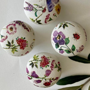 Op de afbeelding: Vier witte ronde ornamenten met bloemen- en vlindermotieven. De ornamenten hebben een witte achtergrond met roze, paarse en groene bloemenpatronen. Een ornament heeft een paarse vlinder met een gele bloem.