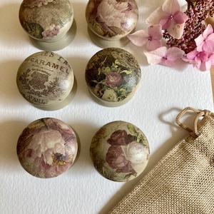 May include: Six decorative knobs with floral and vintage designs. The knobs are made of wood and have a light brown finish. The knobs are arranged in a circle on a white surface. There is a hessian bag in the lower right corner of the image.