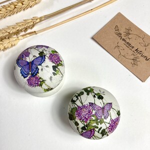 Può includere: Due scatole rotonde bianche con un design floreale con fiori viola e farfalle. Le scatole sono decorate con uno sfondo bianco e un aspetto invecchiato.