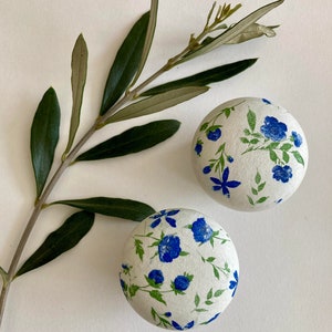 Può includere: Due ornamenti rotondi bianchi con un motivo floreale blu e verde. Gli ornamenti sono su uno sfondo bianco con un ramo di ulivo verde.