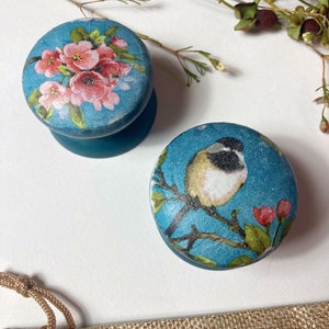 Può includere: Due pomelli rotondi in legno con sfondo blu e disegni floreali. Un pomello presenta un disegno floreale rosa e bianco, mentre l'altro presenta un piccolo uccello appollaiato su un ramo con fiori rossi.