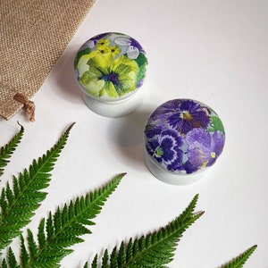 Pode incluir: Dois botões redondos de madeira com desenhos florais. Um botão apresenta violetas amarelas e roxas em um fundo branco. O outro botão apresenta violetas roxas em um fundo branco.