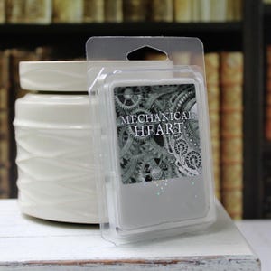May include: A white ceramic wax warmer with a textured design sits next to a clear plastic package of wax melts. The package features the text "MECHANICAL HEART" over a gray and black gear design.