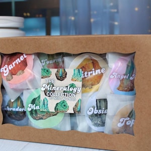 May include: A brown cardboard box with a clear window displaying various wrapped gemstones. Each gemstone is labeled with its name, such as "Garnet" and "Citrine." The box also features a sticker that reads "The Mineralogy Collection."