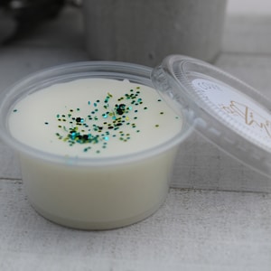 May include: A small, clear plastic container filled with a creamy white wax melt. The wax is sprinkled with teal and black glitter. The lid is partially open, revealing a label with gold text.