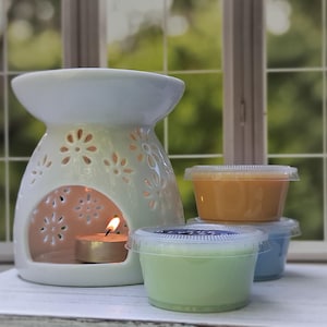 May include: A white ceramic wax warmer with a cut-out floral pattern and a lit tea light candle inside, accompanied by three small, clear plastic containers filled with colored wax melts in shades of light green, blue, and orange.