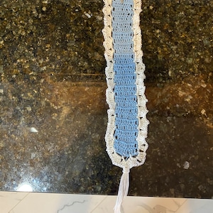 May include: A blue and white crocheted bookmark with a tassel. The bookmark is made with a delicate stitch and has a scalloped edge.