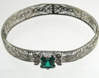 May include: A silver filigree bangle bracelet featuring a large, square, emerald green stone at its centre. The stone is surrounded by smaller, clear stones.