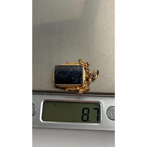 May include: A gold pendant with a rectangular, dark blue stone. The pendant is on a digital scale, which reads 8.7 grams. The pendant has ornate, decorative elements around the stone and a small loop for a chain.
