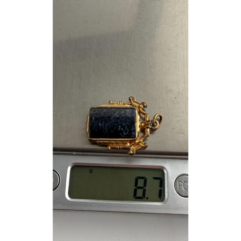 May include: A gold pendant with a rectangular, dark blue stone. The pendant is on a digital scale, which reads 8.7 grams. The pendant has ornate, decorative elements around the stone and a small loop for a chain.