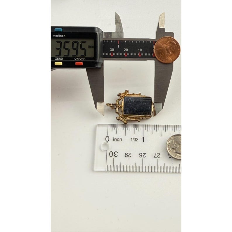 May include: A gold-coloured rectangular pendant with a dark blue stone, measuring 3.6 cm. The pendant is next to a ruler and a penny for size comparison. The pendant has ornate detailing.