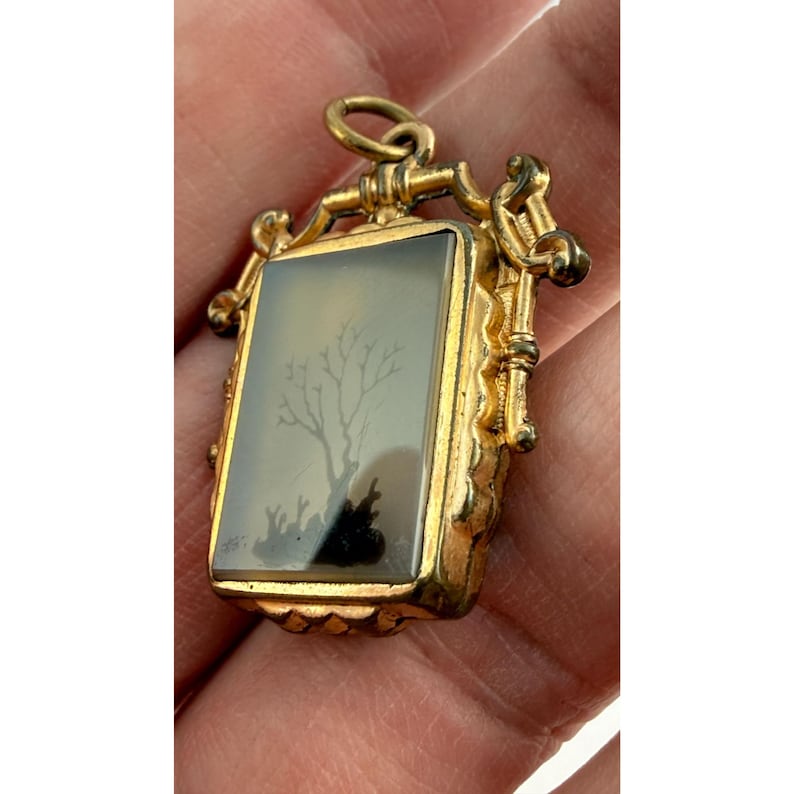 May include: A rectangular gold pendant with a detailed frame. The pendant features a landscape scene with a tree and dark figures. The bail and frame have ornate detailing.