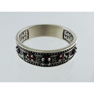 May include: A silver band ring featuring a series of small, round, multi-coloured gemstones including red, green, and black stones set along the outer surface. The interior of the band is engraved with Cyrillic text in a dark, recessed font.
