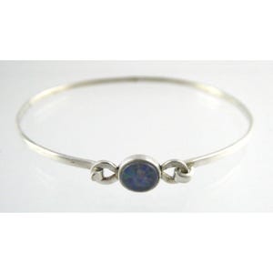 Könnte beinhalten: Ein silbernes Armkettchen mit einem blauen Opal-Edelstein, der in der Mitte eingesetzt ist. Das Armband hat ein schlichtes Design mit zwei Schlaufen auf jeder Seite des Steins.
