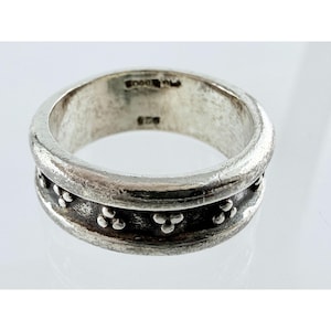 May include: A silver ring with a wide band featuring a recessed center channel decorated with repeating three-dot clusters. The interior of the band is stamped with the hallmark 925, indicating sterling silver composition and a polished finish.