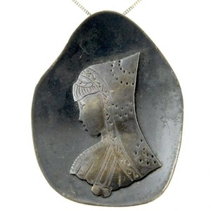 Puede incluir: Un collar de plata con un colgante de piedra negra que presenta una silueta grabada detallada de una mujer con un tocado.