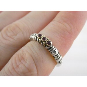 May include: A silver ring with gold accents and three small, round, red gemstones. The ring has a textured design with etched details and is worn on a finger.