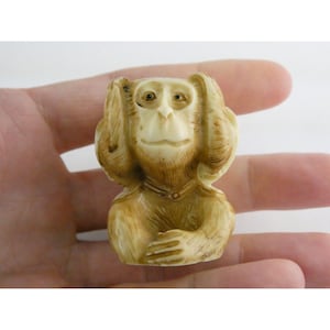 May include: A small, carved monkey figurine in shades of cream and brown. The monkey has its hands over its ears. The figurine is held in a person's hand, with the background out of focus.