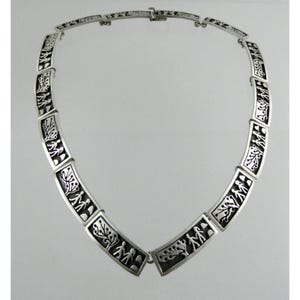 Op de afbeelding: Een zilveren ketting met rechthoekige schakels. Elke schakel toont een zwart, gestileerd ontwerp van bomen en figuren. De ketting, vastgemaakt met een sluiting, wordt gepresenteerd tegen een witte achtergrond. Het ontwerp wordt herhaald.