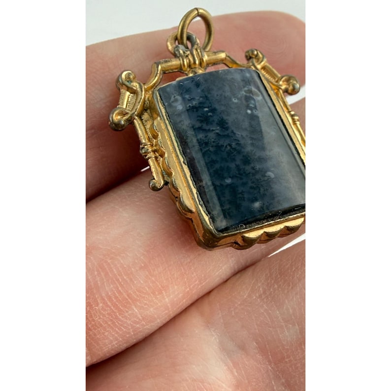May include: A gold-coloured pendant featuring a rectangular, blue-grey stone. The pendant has decorative detailing around the stone and a small loop at the top for a chain. The stone has a marbled appearance.