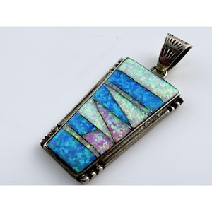 May include: A rectangular silver pendant with a mosaic of blue, white, and purple opal stones. The pendant has a decorative bail and a beaded border. The opal stones are arranged in a geometric pattern.