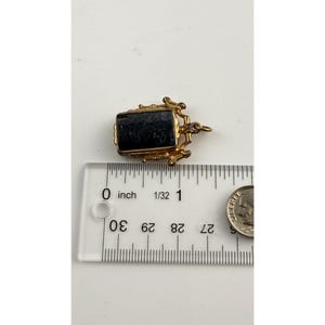 May include: A small, rectangular pendant with a dark blue stone set in a gold-coloured metal frame. The pendant is approximately 1.9 cm long. A ruler and a coin are next to the pendant.