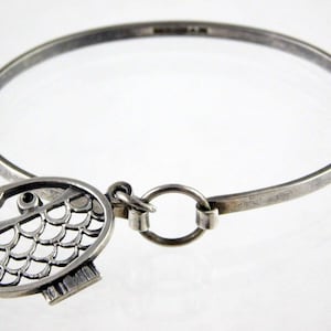 Könnte beinhalten: Ein silbernes Armreif-Armband mit einem Anhänger in Form eines Fisches mit einem Ausschnittmuster. Der Anhänger ist mit einem silbernen Ring am Armband befestigt.