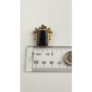 May include: A small, rectangular pendant with a dark stone set in a gold-coloured metal frame. The pendant is approximately 1.9 cm long. The frame has ornate detailing, and the pendant is shown with a ruler and a coin for scale.