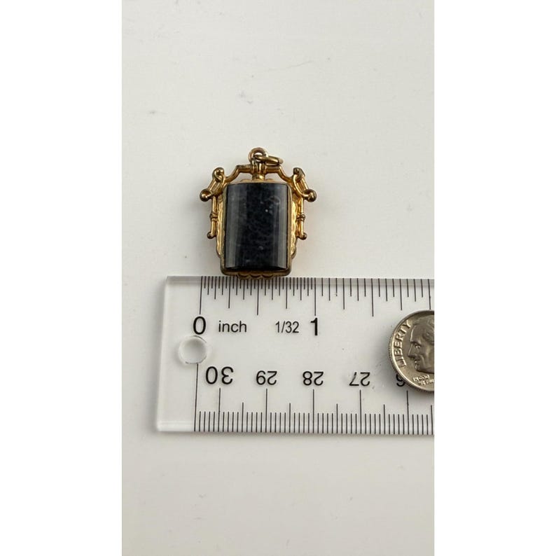 May include: A small, rectangular pendant with a dark stone set in a gold-coloured metal frame. The pendant is approximately 1.9 cm long. The frame has ornate detailing, and the pendant is shown with a ruler and a coin for scale.