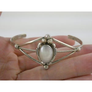 May include: Silver cuff bracelet featuring an oval, pearlescent centerpiece. The bracelet has a geometric design with decorative floral accents and twisted rope detailing. A classic piece of jewelry.