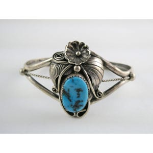May include: A silver cuff bracelet with a central oval turquoise stone. The bracelet features a floral design with leaves and a flower at the top. The bracelet has a delicate chain detail on each side.