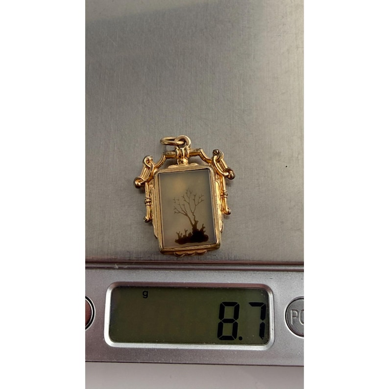 May include: A gold pendant with a rectangular, framed design. The pendant features a landscape scene with a tree and figures. The pendant weighs 8.7 grams. The pendant is on a digital scale.